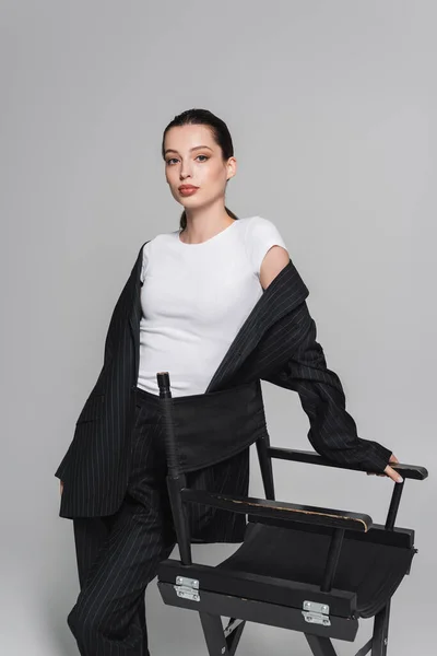 Mulher na moda em terno olhando para a câmera perto de cadeira dobrável no fundo cinza — Fotografia de Stock