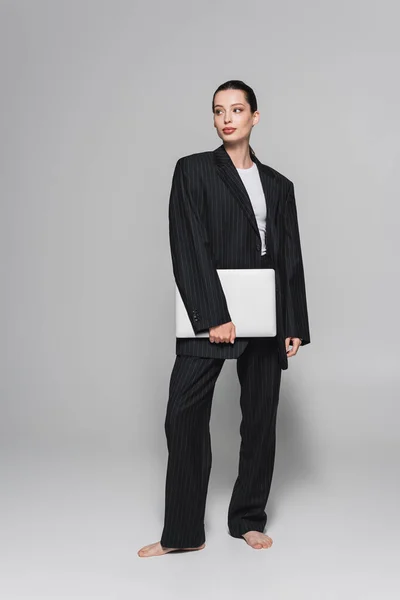 Стильная босиком женщина в костюме держит ноутбук и смотрит в сторону на сером фоне — стоковое фото