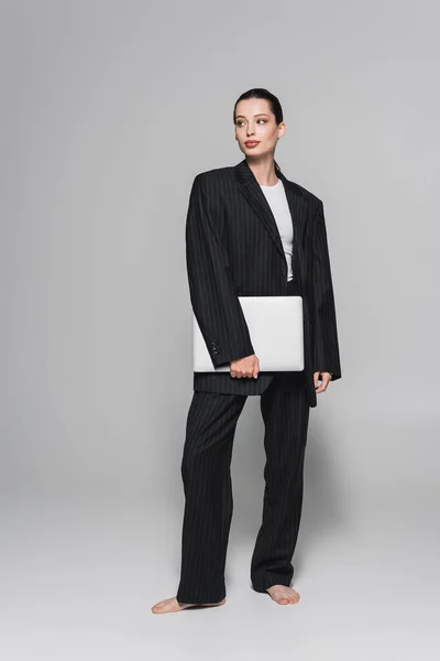 Stylish barefoot woman in suit holding laptop and looking away on grey background — Stock Photo
