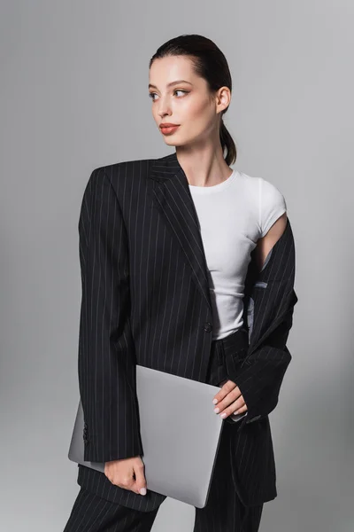 Hübsche brünette Frau im gestreiften Anzug hält Laptop isoliert auf grau — Stockfoto