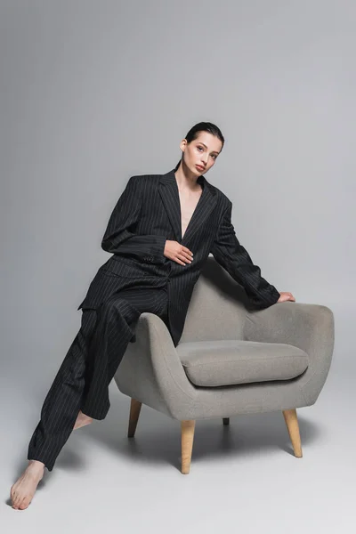 Pleine longueur de jeune femme à la mode en costume posant près du fauteuil sur fond gris — Photo de stock