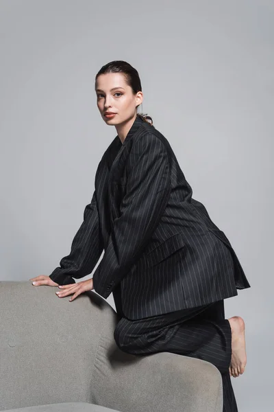 Brünette Frau im gestreiften Anzug posiert in der Nähe von Sessel isoliert auf grau — Stockfoto