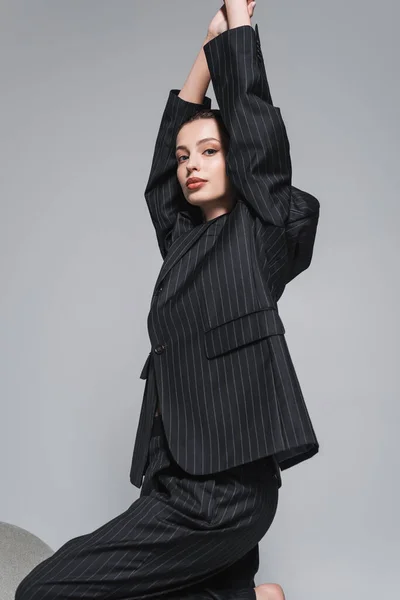 Modelo de moda en chaqueta a rayas y pantalones posando cerca del sillón y mirando a la cámara aislada en gris - foto de stock