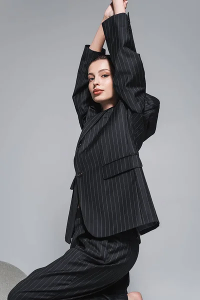 Fashionable model in striped jacket and pants posing near armchair and looking at camera isolated on grey — Stock Photo