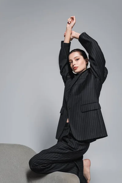 Модель босоніж у смугастому костюмі, що піднімає руки біля крісла ізольовано на сірому — стокове фото