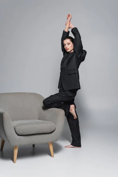 Модная и босоногая модель в костюме, смотрящая на камеру возле кресла на сером фоне — стоковое фото
