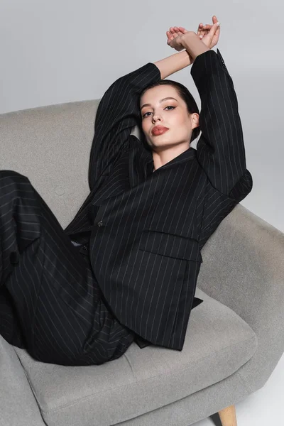 Stilvolle Frau im gestreiften Anzug blickt in die Kamera, während sie auf einem Sessel auf grauem Hintergrund sitzt — Stockfoto