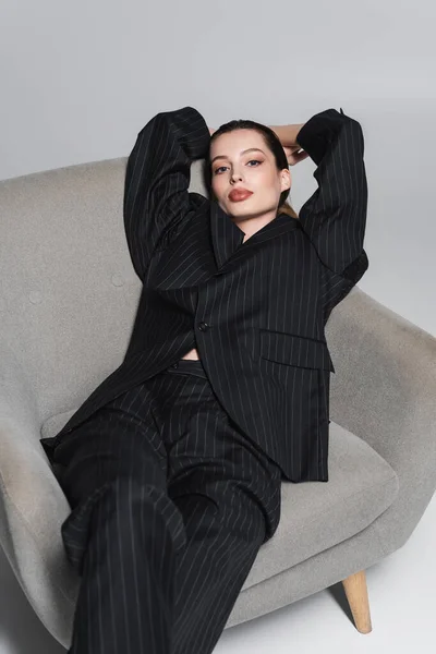 Jeune femme tendance en costume rayé noir regardant caméra sur fauteuil sur fond gris — Photo de stock