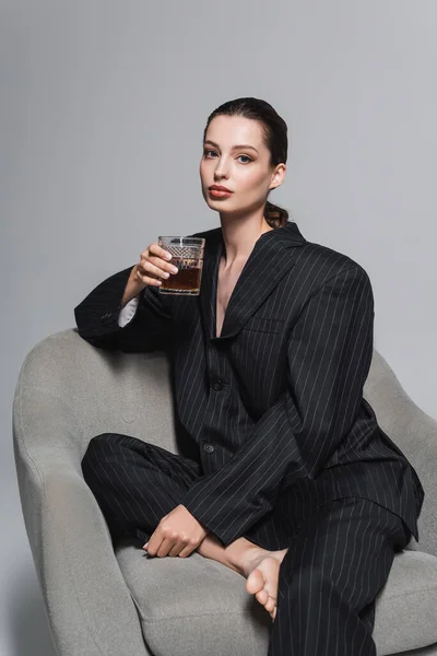 Stylish model in black suit holding glass of whiskey while sitting on armchair on grey background — Stock Photo