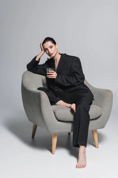 Barefoot woman in black suit holding glass of whiskey while sitting on armchair on grey background — Stock Photo