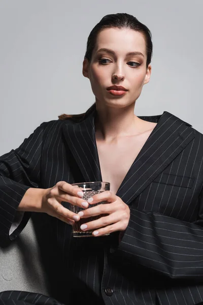 Portrait of woman in striped jacket holding glass of whiskey while sitting on armchair isolated on grey — Stock Photo