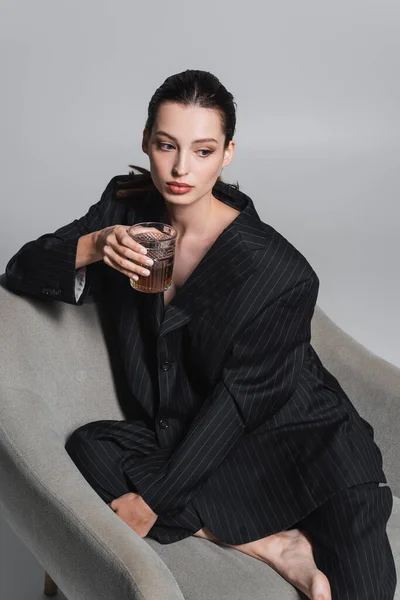 Brunette woman in striped suit holding whiskey while sitting on armchair on grey background — Stock Photo