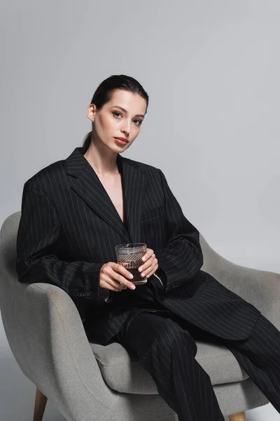 Stylish woman in striped suit holding whiskey and looking at camera on armchair on grey background — Stock Photo