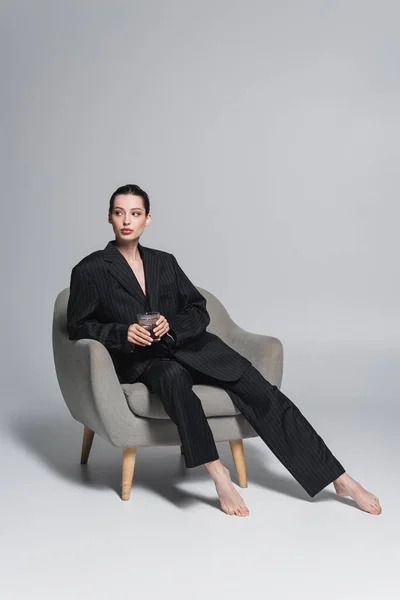 Barfüßige Frau im schwarzen Anzug mit Whiskeyglas auf Sessel vor grauem Hintergrund — Stockfoto