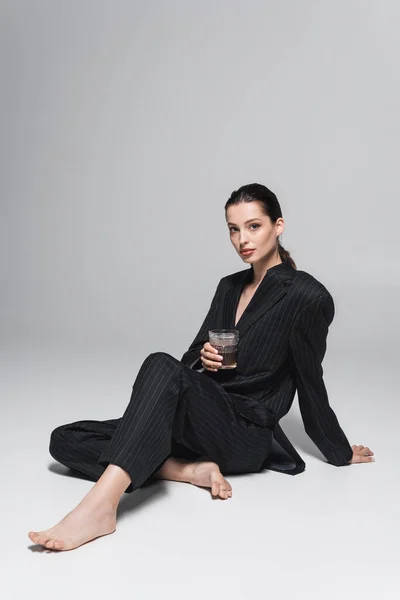 Модель босоніж в чорному костюмі тримає келих віскі, сидячи на сірому фоні — стокове фото