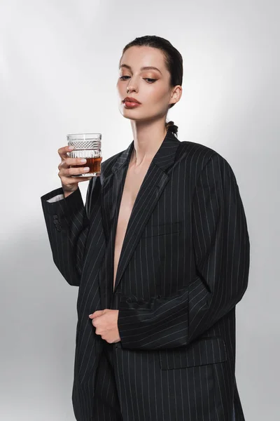 Stylish young woman in striped jacket holding whiskey on abstract grey background — Stock Photo