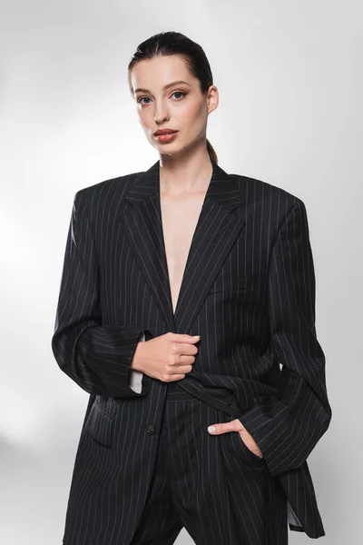 Портрет модной модели в костюме, держащей руку в кармане на сером фоне — стоковое фото
