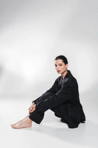 Jovem modelo descalço em terno listrado olhando para a câmera enquanto sentado em fundo cinza — Fotografia de Stock