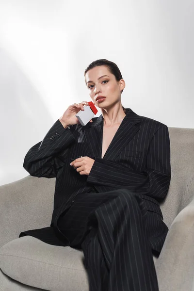 Mujer de moda en traje de rayas que sostiene la caja de cigarrillos mientras está sentado en el sillón sobre fondo gris - foto de stock