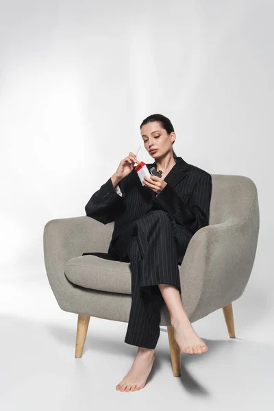 Barefoot model in suit holding cigarette and lighter while sitting on armchair on grey background — Stock Photo