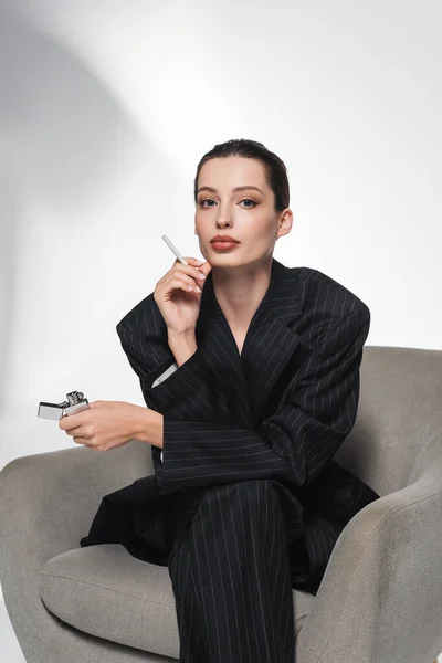 Модная женщина в полосатом костюме держит сигарету и зажигалку, сидя на кресле на абстрактном сером фоне — стоковое фото