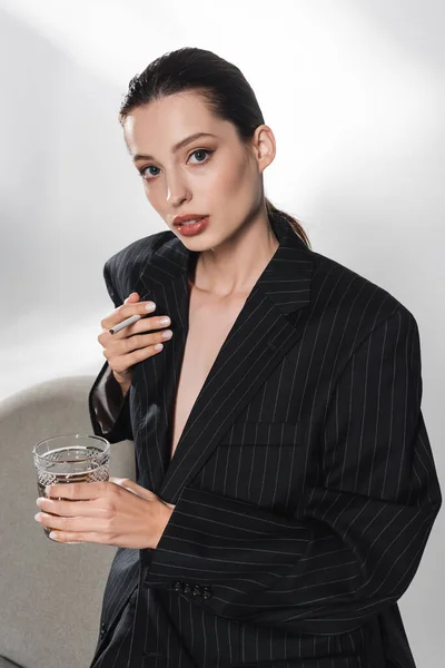 Stylish woman in black jacket holding glass of whiskey and cigarette near armchair on grey background — Stock Photo