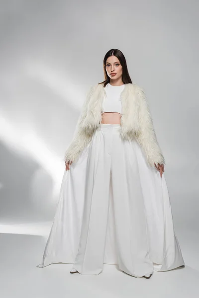 Полная длина брюнетки женщина в искусственной меховой куртке и белой одежде позирует на абстрактном сером фоне — стоковое фото
