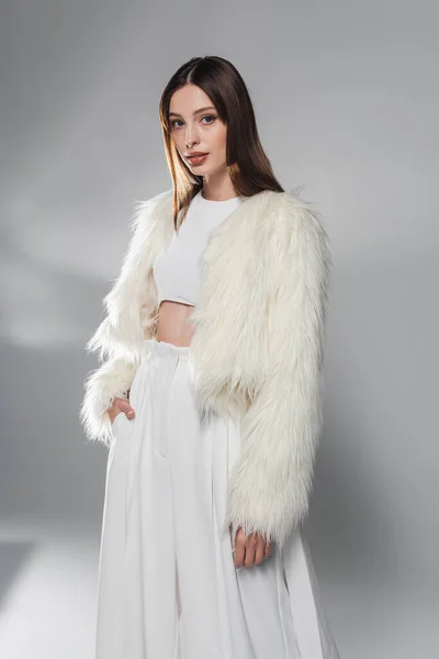 Модная женщина в искусственном меховом пиджаке и белой одежде позирует на сером фоне — стоковое фото