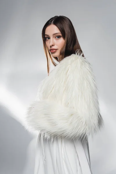 Stylish brunette woman in faux fur jacket looking at camera on abstract grey background — Stock Photo