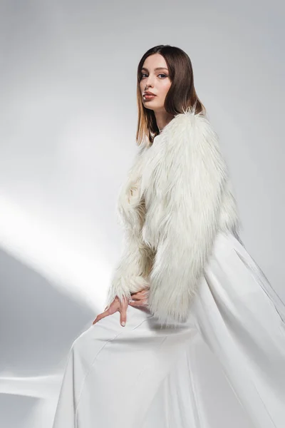 Модная модель в белой одежде и искусственной меховой куртке, смотрящая в камеру на абстрактном сером фоне — стоковое фото