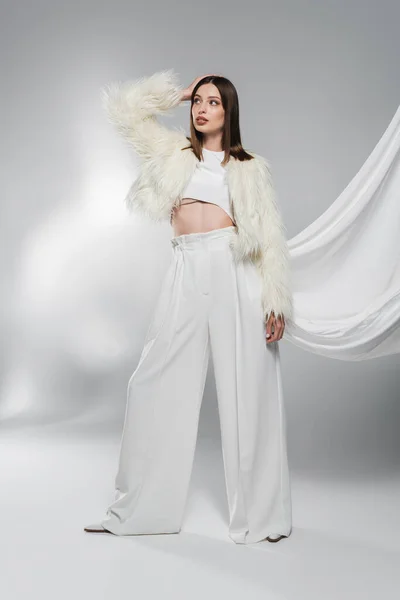 Mulher na moda em casaco de pele falso e calças brancas posando no fundo cinza abstrato — Fotografia de Stock