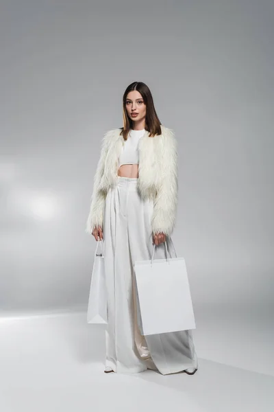 Volle Länge der trendigen Frau in weißer Kleidung mit Einkaufstaschen auf abstrakt grauem Hintergrund — Stockfoto