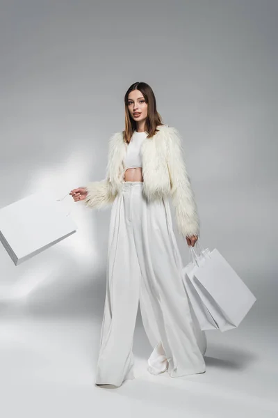 Donna alla moda in pelliccia sintetica giacca e pantaloni che tengono borse della spesa su sfondo grigio astratto — Foto stock