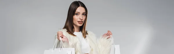 Stylish model in white faux fur jacket holding shopping bags on grey background, banner — Stock Photo