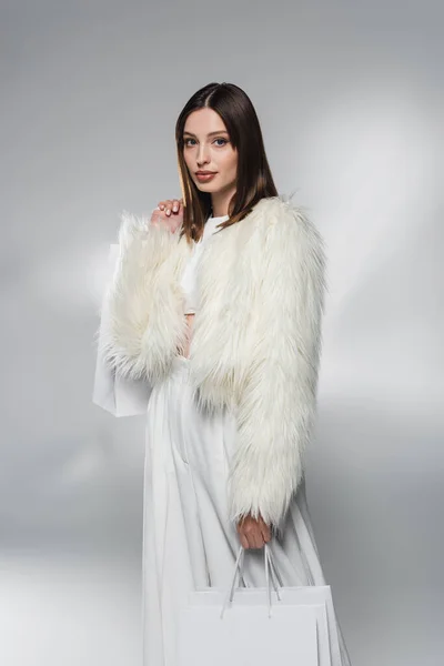 Модная брюнетка в белом искусственном меховом пиджаке держит сумки для покупок на абстрактном сером фоне — стоковое фото