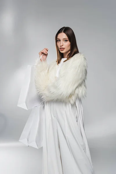 Fashionable woman in white faux fur jacket holding shopping bags and looking at camera on abstract grey background — Stock Photo