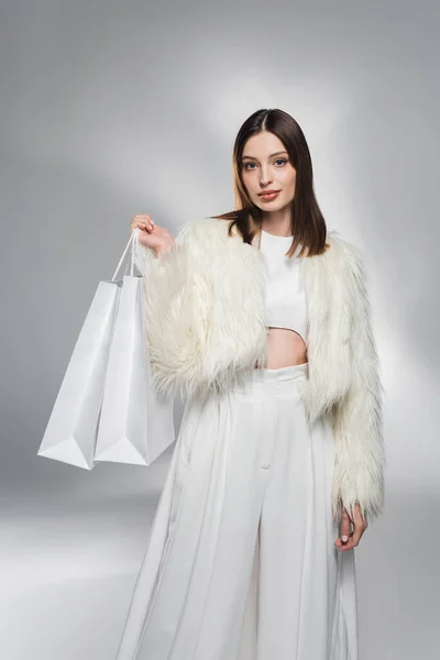 Модна жінка в штучній куртці, що тримає білі сумки для покупок на абстрактному сірому фоні — стокове фото