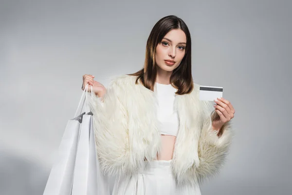 Jeune femme élégante veste en fausse fourrure tenant carte de crédit et sacs à provisions sur fond gris abstrait — Photo de stock