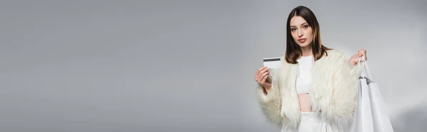 Giovane donna in giacca di pelliccia ecologica con shopping bag bianchi e carta di credito su sfondo grigio, banner — Foto stock