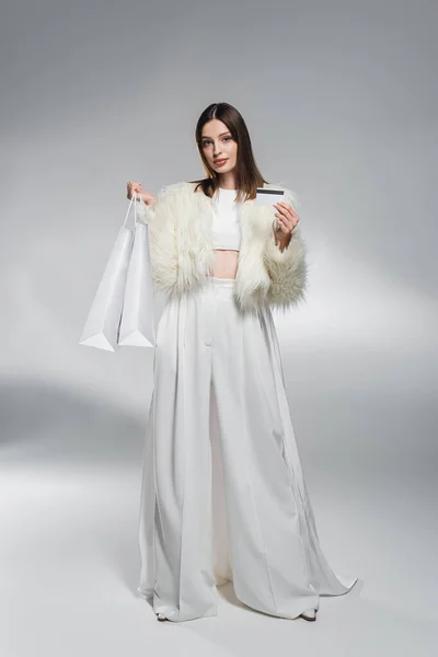 Полная длина модной женщины в белой одежде, держащей кредитки и сумки для покупок на абстрактном сером фоне — стоковое фото
