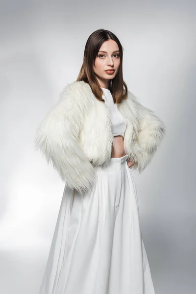 Модная женщина в белой одежде и искусственной меховой куртке, держащая руки на бедрах на абстрактном сером фоне — стоковое фото