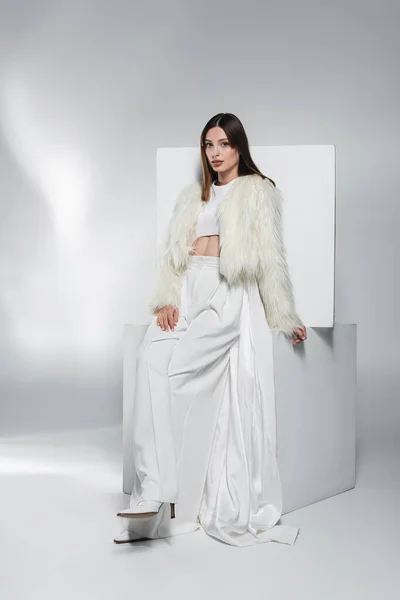 Elegante giovane donna in abiti bianchi e giacca di pelliccia ecologica in posa vicino a cubi su sfondo grigio astratto — Foto stock