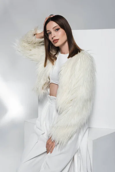 Trendy brunette woman in white faux fur jacket posing near cubes on abstract grey background — Stock Photo
