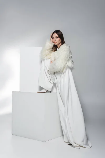 Elegante donna in abiti bianchi e giacca di pelliccia ecologica guardando la fotocamera vicino a cubi su sfondo grigio astratto — Foto stock