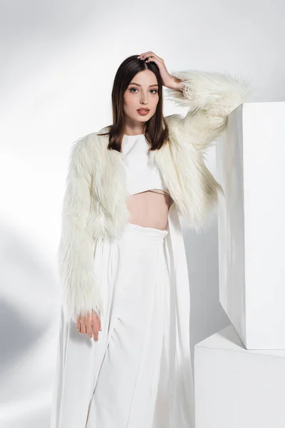 Pretty young woman in white faux fur jacket and crop top leaning on cube while posing on grey — Stock Photo