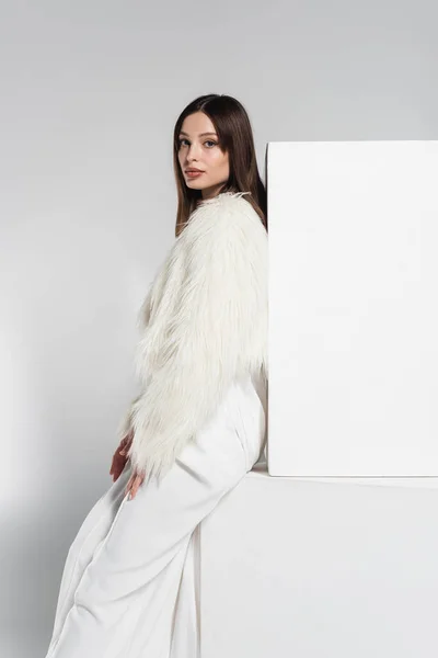 Pretty young woman in white faux fur jacket leaning on cubes while looking at camera on grey — Stock Photo