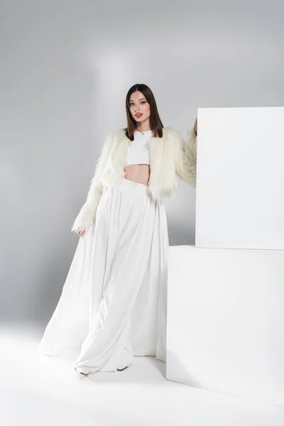 Longitud completa de la mujer joven en chaqueta de piel sintética y traje blanco total apoyado en cubos mientras posando en gris - foto de stock