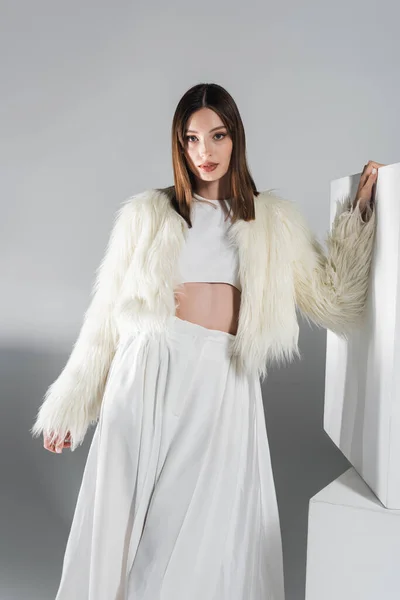 Giovane donna in giacca di pelliccia ecologica e total outfit bianco in posa vicino a cubi sul grigio — Foto stock