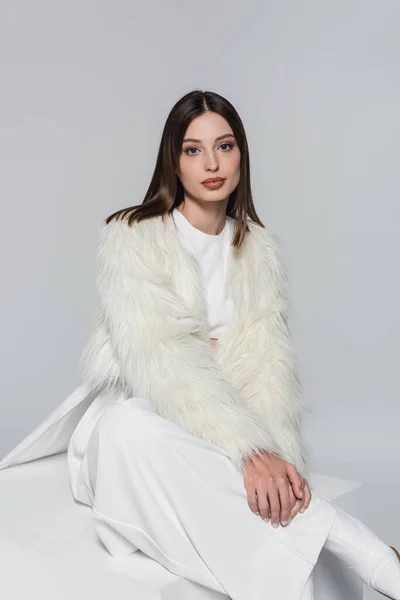 Young woman in white faux fur jacket and total white outfit sitting on cube isolated on grey — Stock Photo