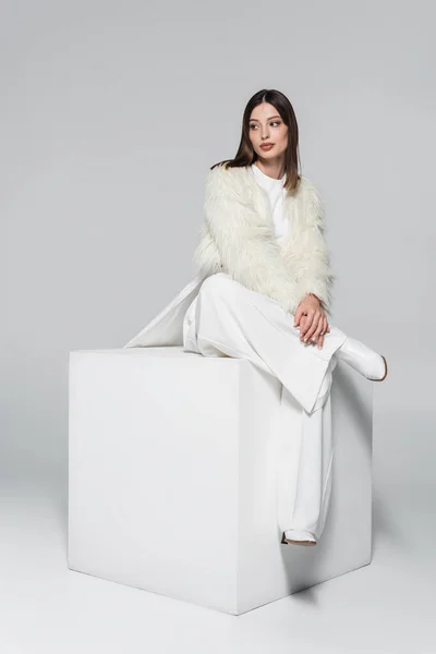 Longitud completa de la mujer joven en chaqueta de piel sintética y traje blanco total sentado en cubo en gris - foto de stock